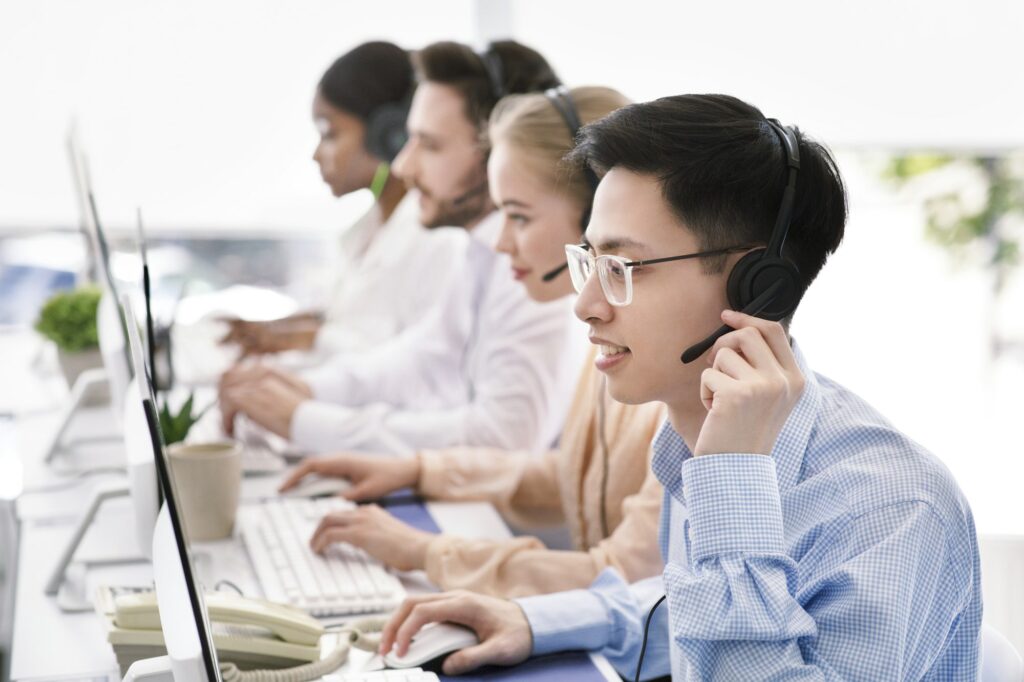 Asian helpdesk representative in headphones with his colleagues providing tech support services to